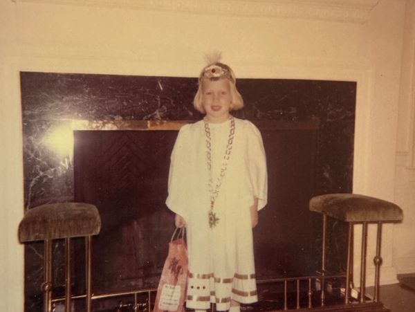 A young girl in a white dress and headband stands by a fireplace holding a bag.